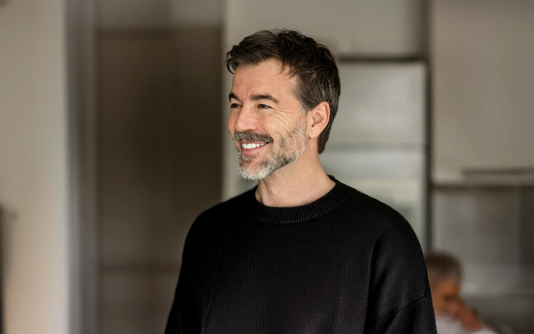 A middle-aged man with salt-and-pepper hair and a groomed beard, smiling and looking off-camera. He is wearing a black textured crewneck sweater in a brightly lit indoor setting.