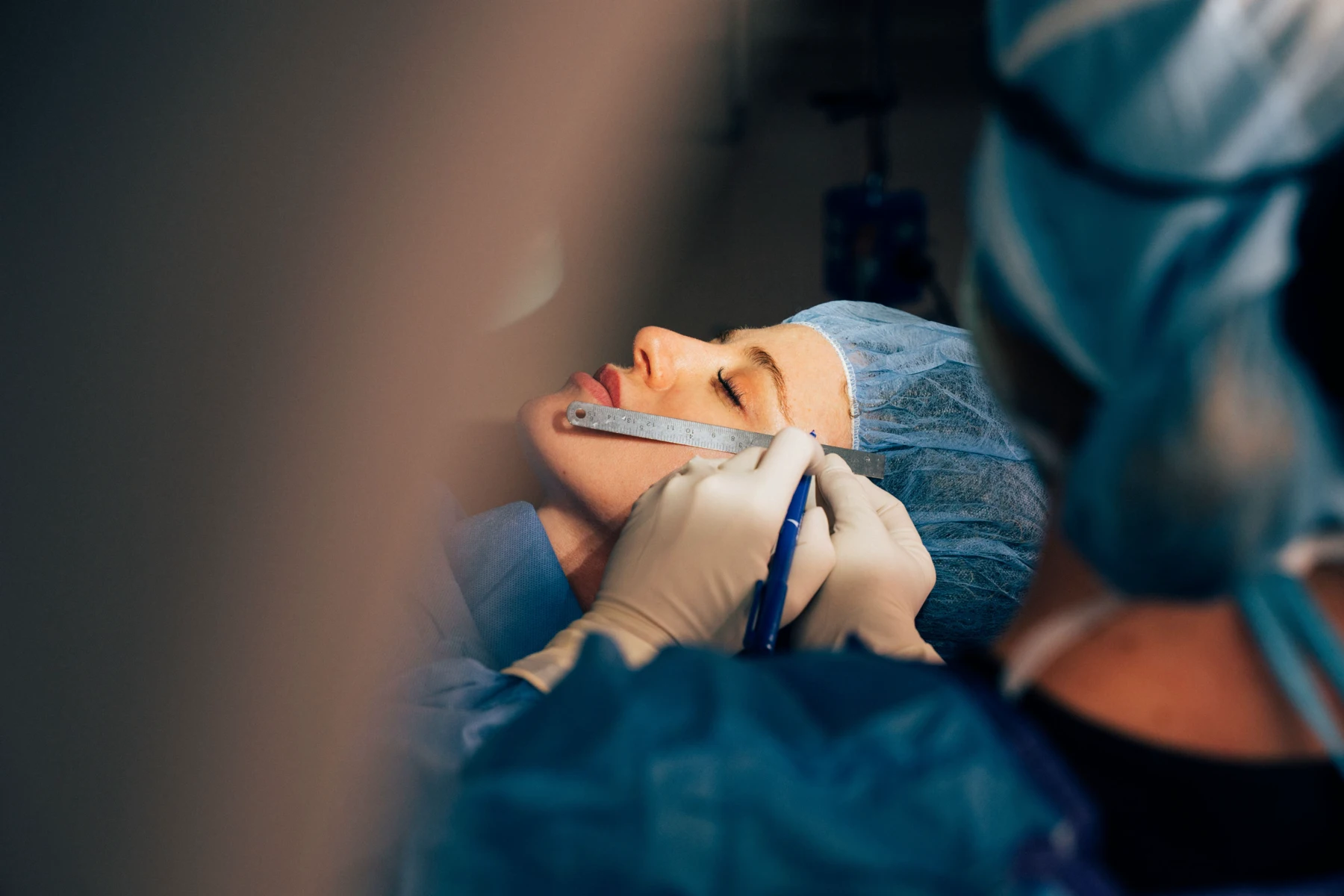 A close-up of a patient in a blue surgical cap undergoing a facial procedure. A medical professional uses a metal ruler and a blue marker to precisely measure and mark the skin.