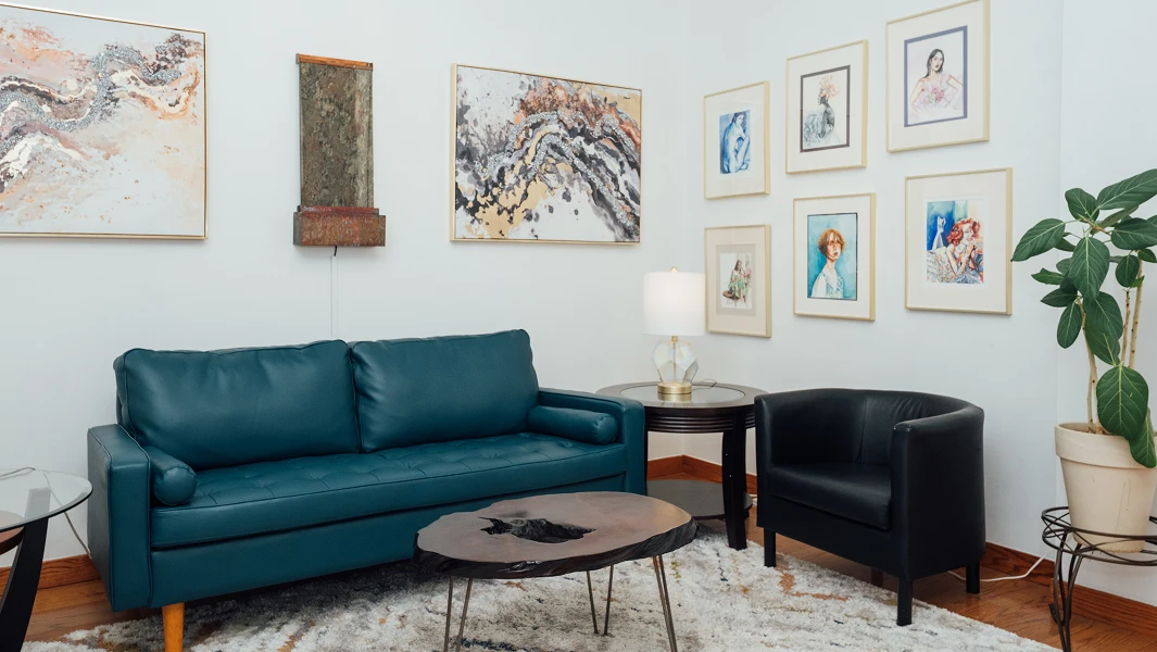 A chic and inviting patient waiting area featuring a deep teal leather sofa, a natural wood coffee table, and a gallery wall of colorful fine art portraits.