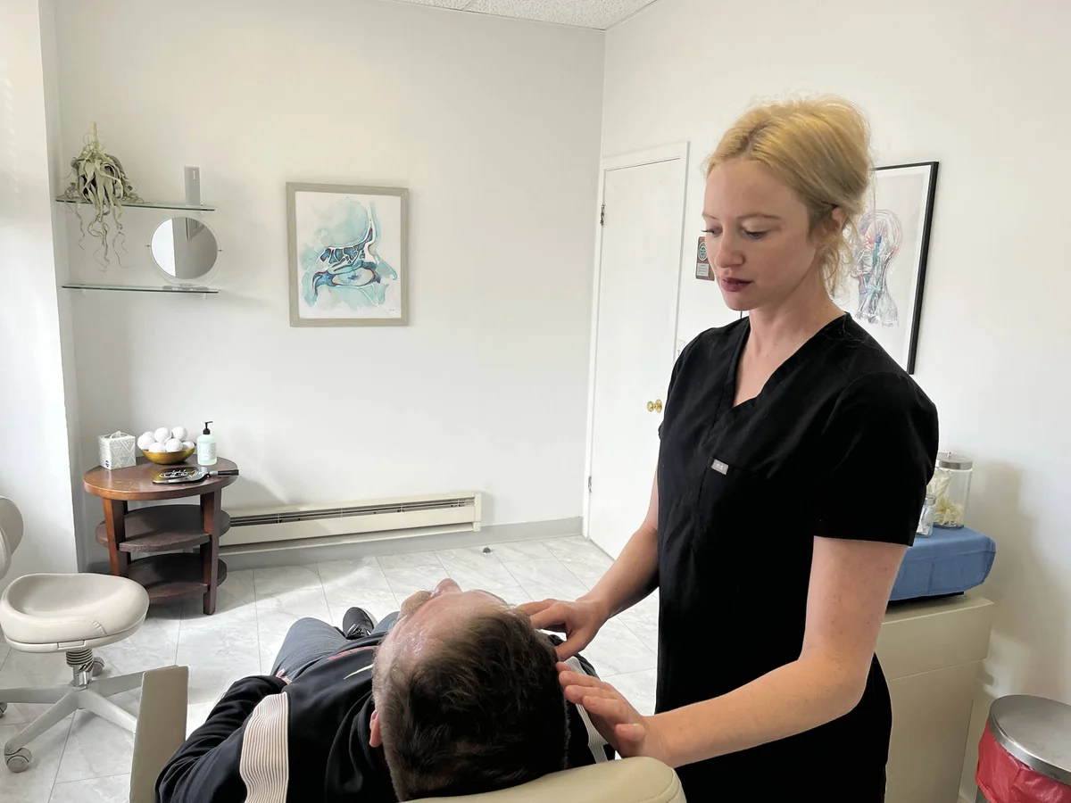 Dr. Meaghan Noud, wearing black scrubs, performs a gentle facial assessment on a patient in a bright, modern clinical exam room.