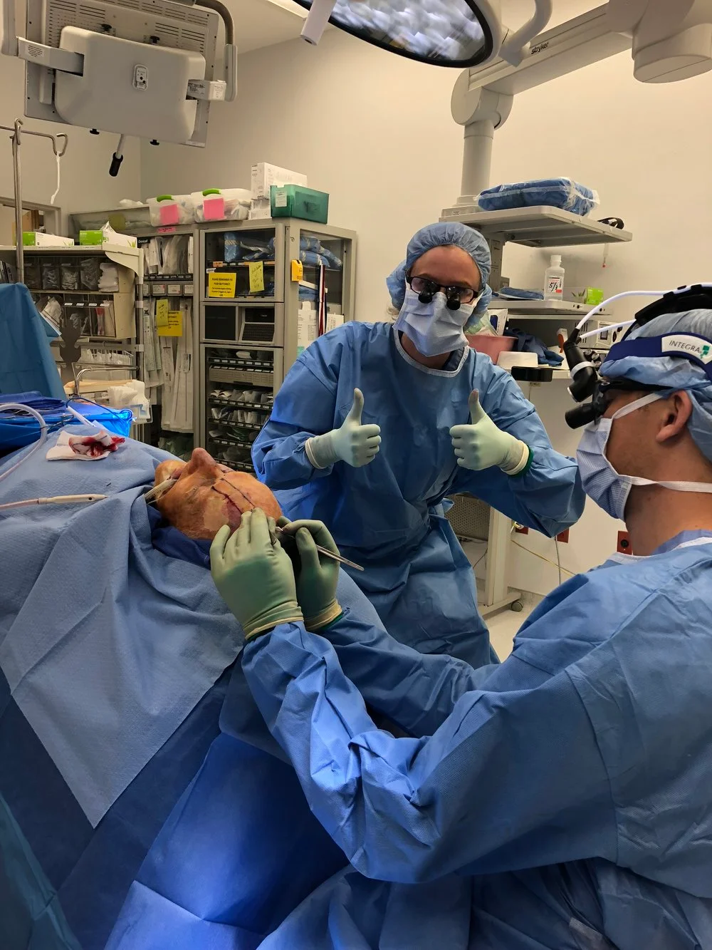 Dr. Noud and a colleague in full sterile surgical blue scrubs giving a "thumbs up" during a procedure in a high-tech operating suite.