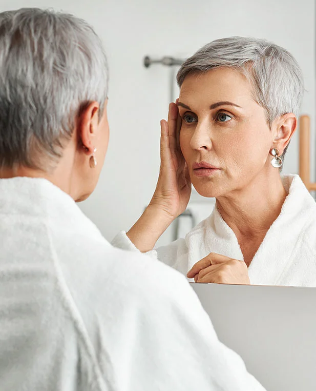 An older woman in a white robe looking in a mirror and touching her face, showcasing the results of skin tightening and resurfacing treatments.