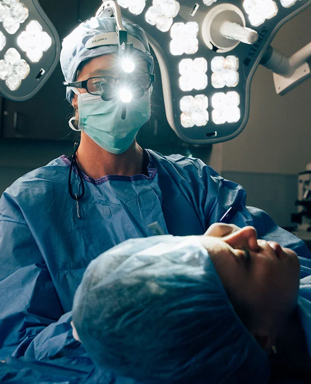 Dr. Noud performs a deep plane facelift in a sterile operating room. She is wearing surgical magnification loupes and a headlight, focusing intently on the patient's facial contours.