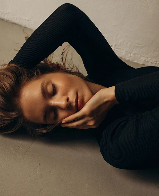 A woman lying down with her eyes closed and hand to her face, wearing a black ribbed top, emphasizing defined cheekbones and facial contours.