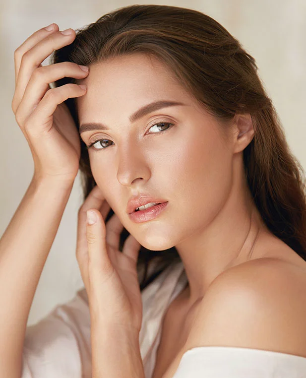 Close up of a young woman with shoulder-length brown hair gazes softly at the camera, her hands gently resting on her head and face. She wears a loose, off-white, off-the-shoulder top. Her smooth, light-toned skin glows under soft, diffused lighting. The muted beige background is softly blurred, keeping the focus on her serene expression and natural beauty.
