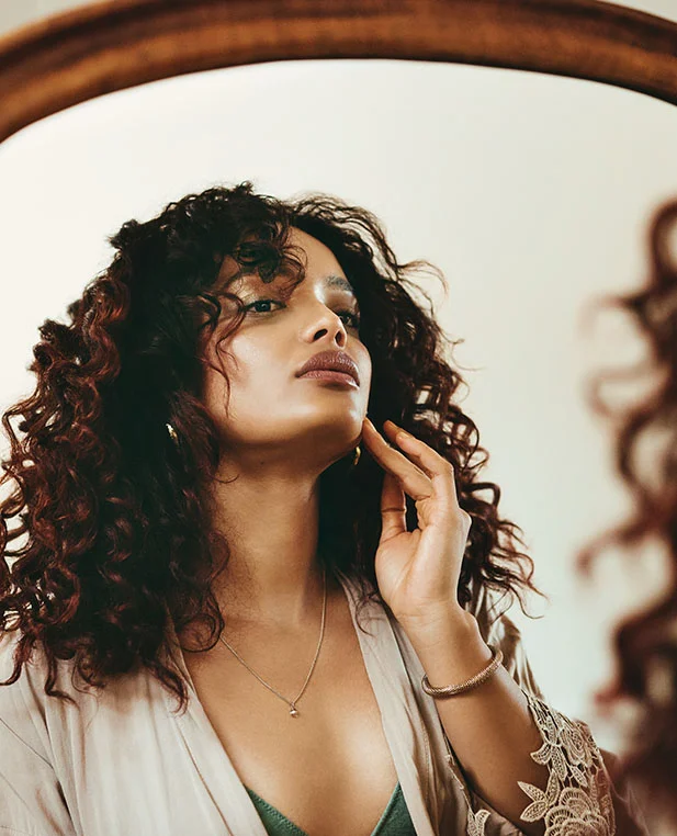 A young woman with long, curly brown hair, wearing a beige robe with lace trim. She is looking at her reflection in a mirror and touching her chin with her right hand. She is wearing a thin gold necklace and a beaded bracelet.