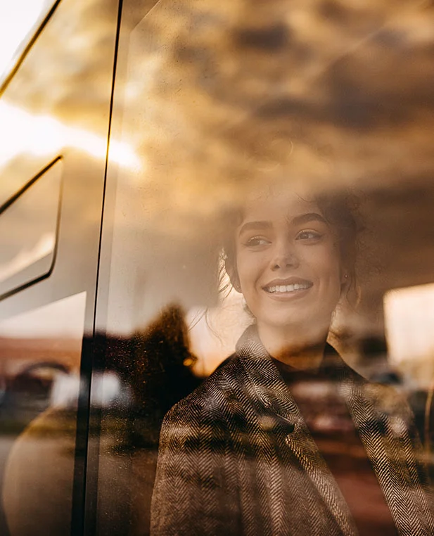 out-of-town-intro.webp A woman looks out of a window at a sunset, her reflection suggesting a journey to receive specialized care from Dr. Noud.