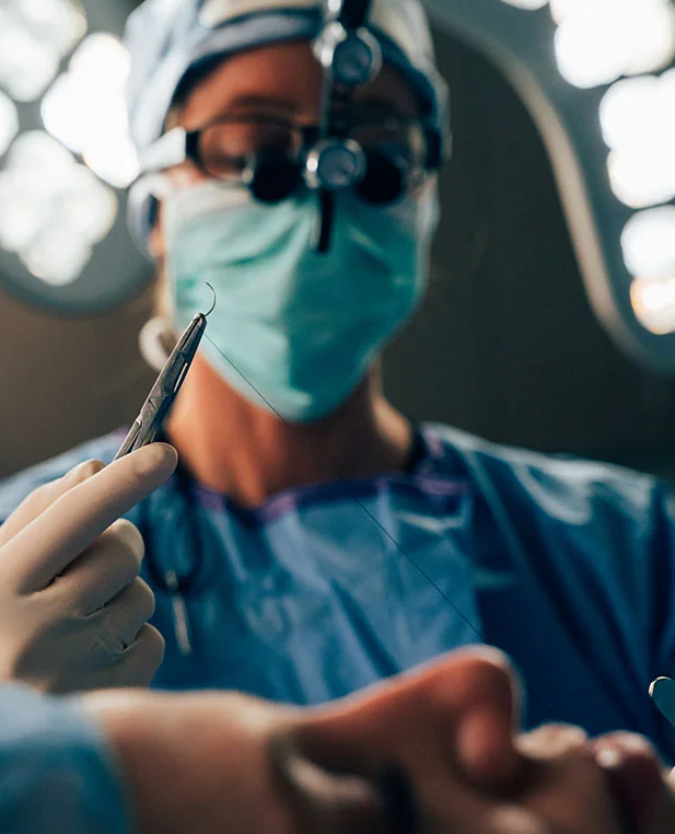 Dr. Noud, wearing surgical loupes and a mask, holds a curved surgical needle and suture during a revision rhinoplasty procedure.