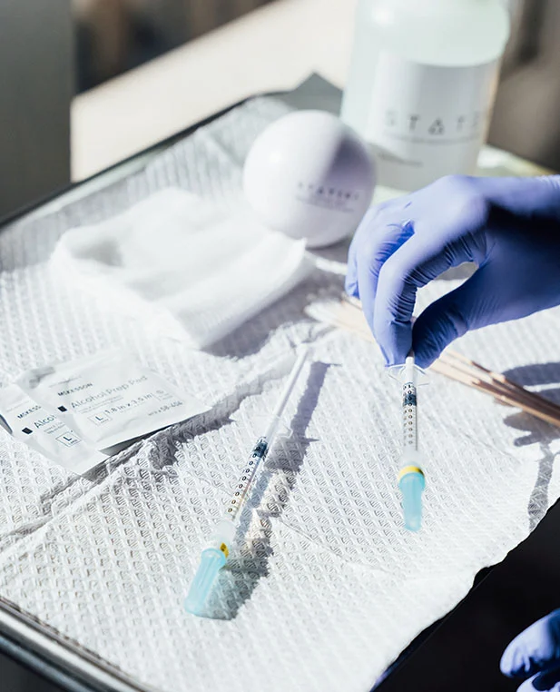A clinical tray set up for a Sculptra treatment at Statera. A gloved hand reaches for one of two prepared syringes resting on a sterile drape next to alcohol prep pads and branded clinic materials.